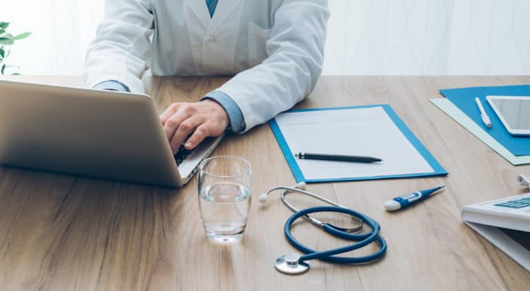 Medical professional at a desk with a glass of water, stethoscope, thermometer, laptop, and tablet