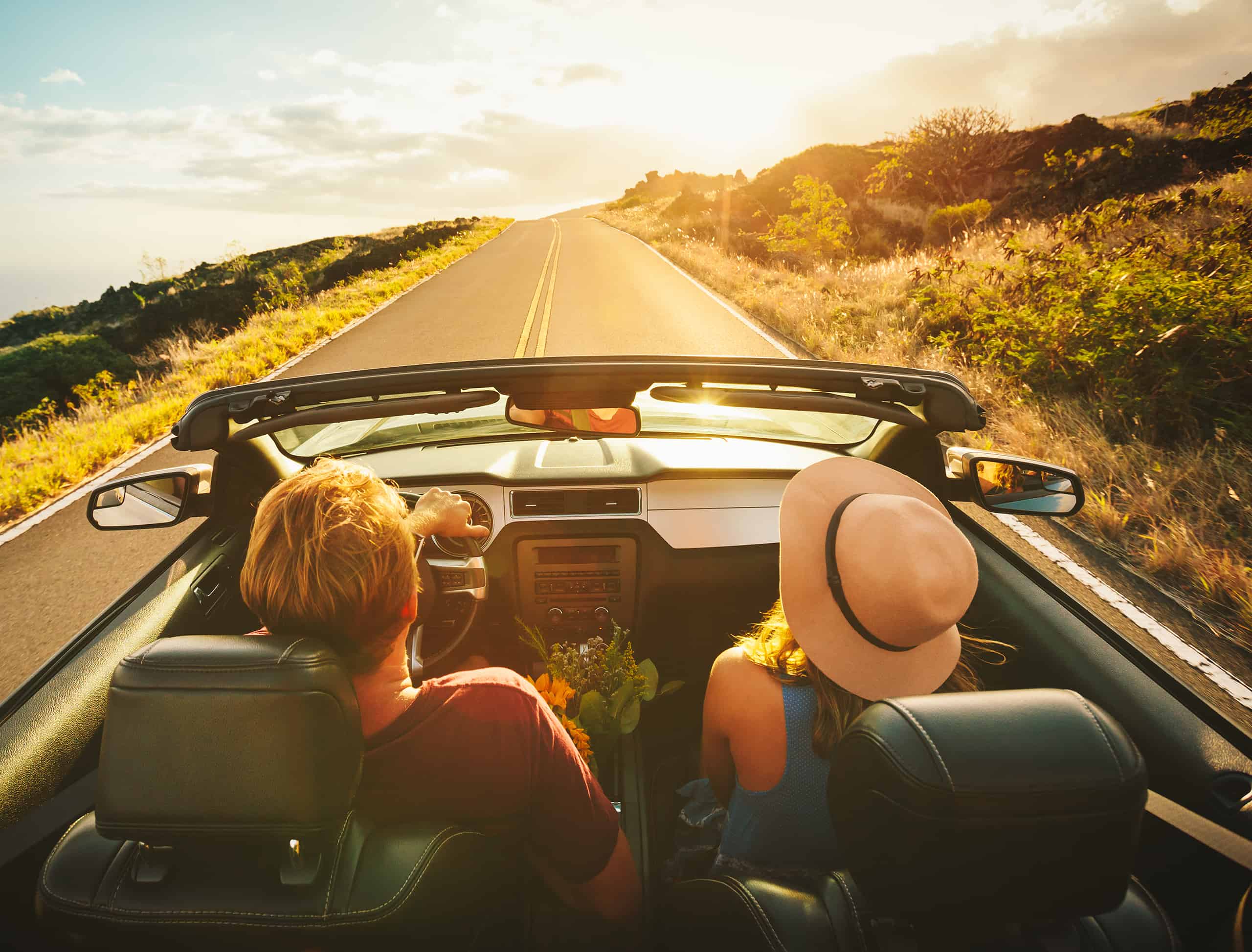 Young couple in convertible driving down road toward sunset